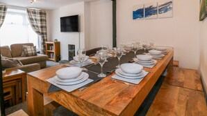 A long wooden dining table set with white plates, glasses, and cutlery, flanked by a sofa and a television. - Fistral Quay (Newquay)