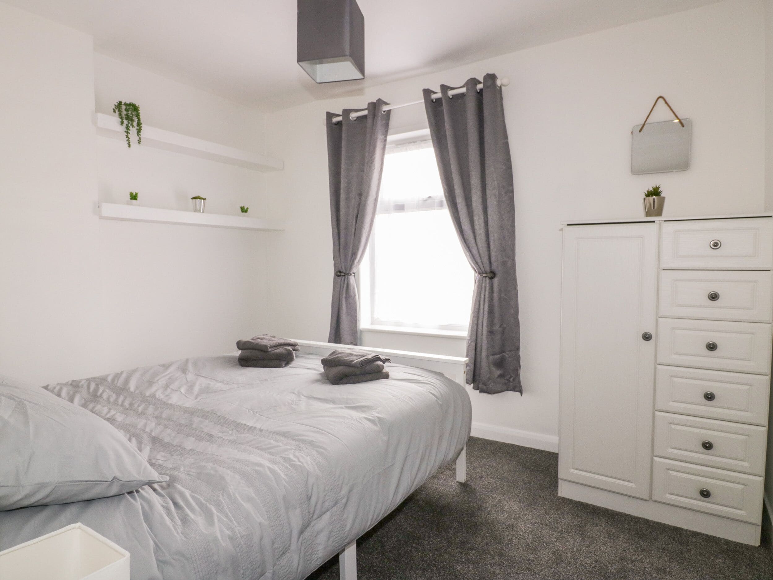 A bedroom with a bed, a dresser, a window with curtains, and a shelf with plants.
