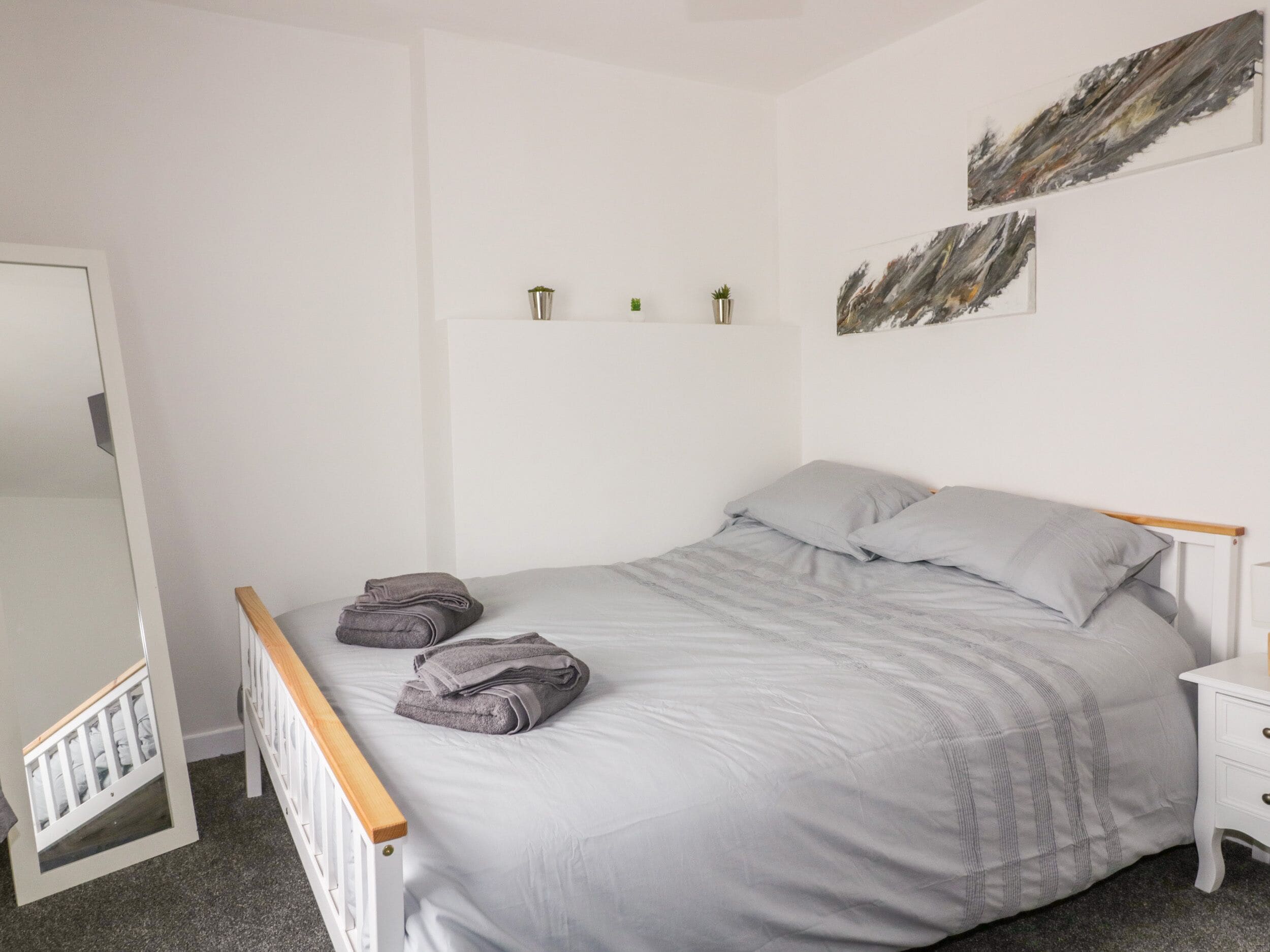 A neatly made bed with grey linens and pillows, a white bedside table with a drawer, and a small plant on the wall.