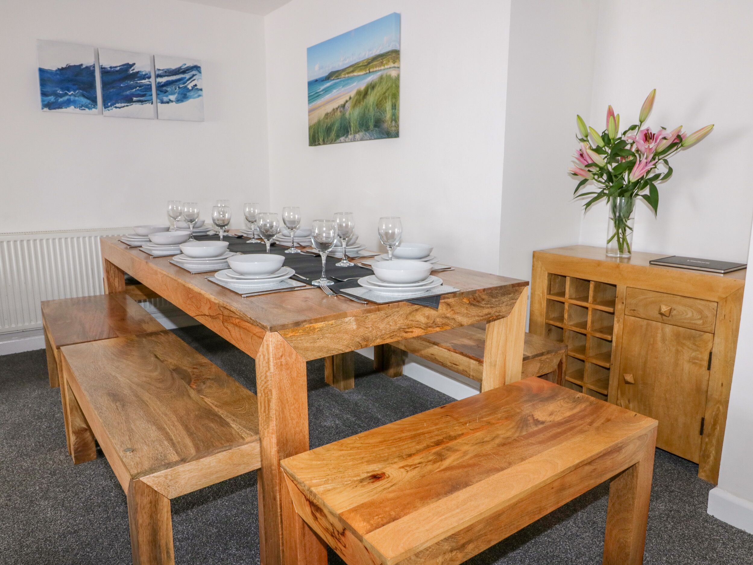 A wooden dining table set with plates, glasses, and cutlery, flanked by benches, with artwork on the walls and a vase with flowers on a cabinet.