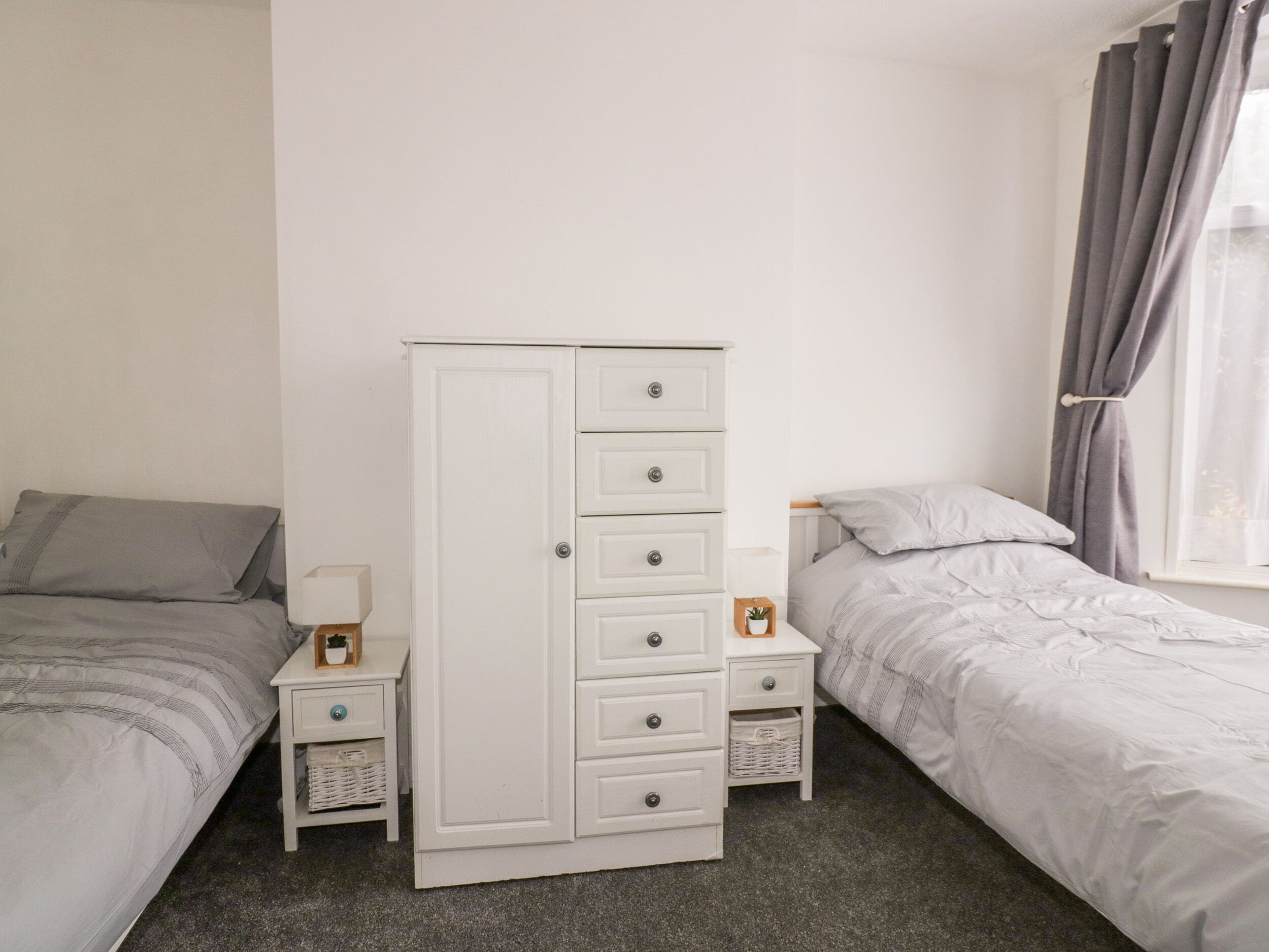 A bedroom with two beds, a white wardrobe, bedside tables with lamps, and a window with curtains.