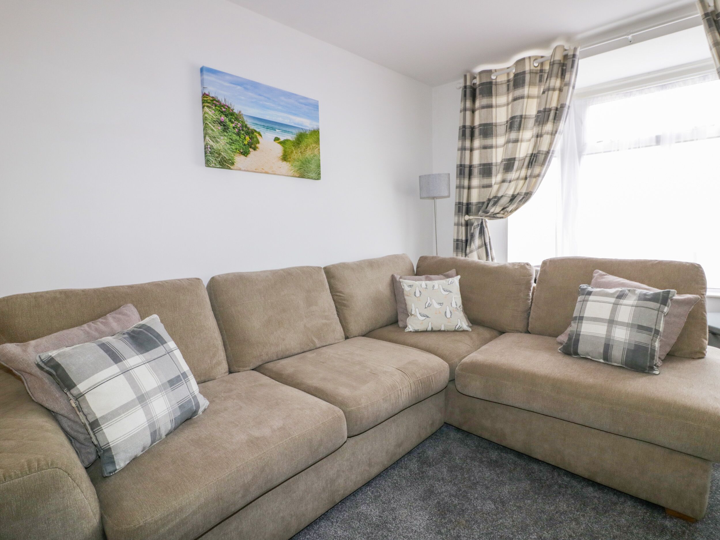 A beige sectional sofa with plaid cushions, a painting of a beach scene, a floor lamp, and a window with plaid curtains.