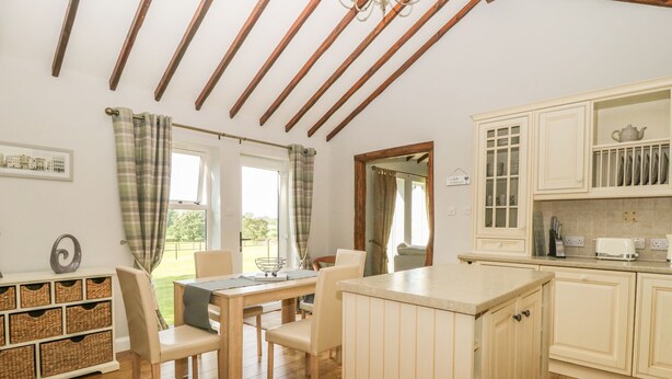 A cozy kitchen with wooden beams, a dining area with a table and chairs, and a view of the outdoors through large windows. - The Barn at Orchard Farm (York)