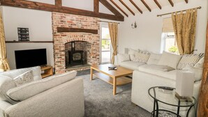 A cozy living room with a brick fireplace, wooden beams, a sofa, a coffee table, and a TV. - The Barn at Orchard Farm (York)