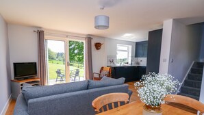 A living room with a grey sofa, a wooden dining table with a vase of white flowers, a TV, and a view of a garden through a sliding glass door. - Owl Cottage (Llandrindod Wells)