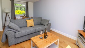 A living room with a grey sofa, a wooden coffee table with a wine bottle and glass, a lamp, and a window with curtains. - Owl Cottage (Llandrindod Wells)