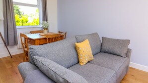 A grey sofa with a yellow pillow, a wooden dining table with chairs, and a window with curtains. - Owl Cottage (Llandrindod Wells)