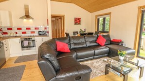 A modern living room with a sectional sofa, a glass coffee table, and a kitchen area with white cabinets and red accents. - Chale Farm Lodge (Ventnor)