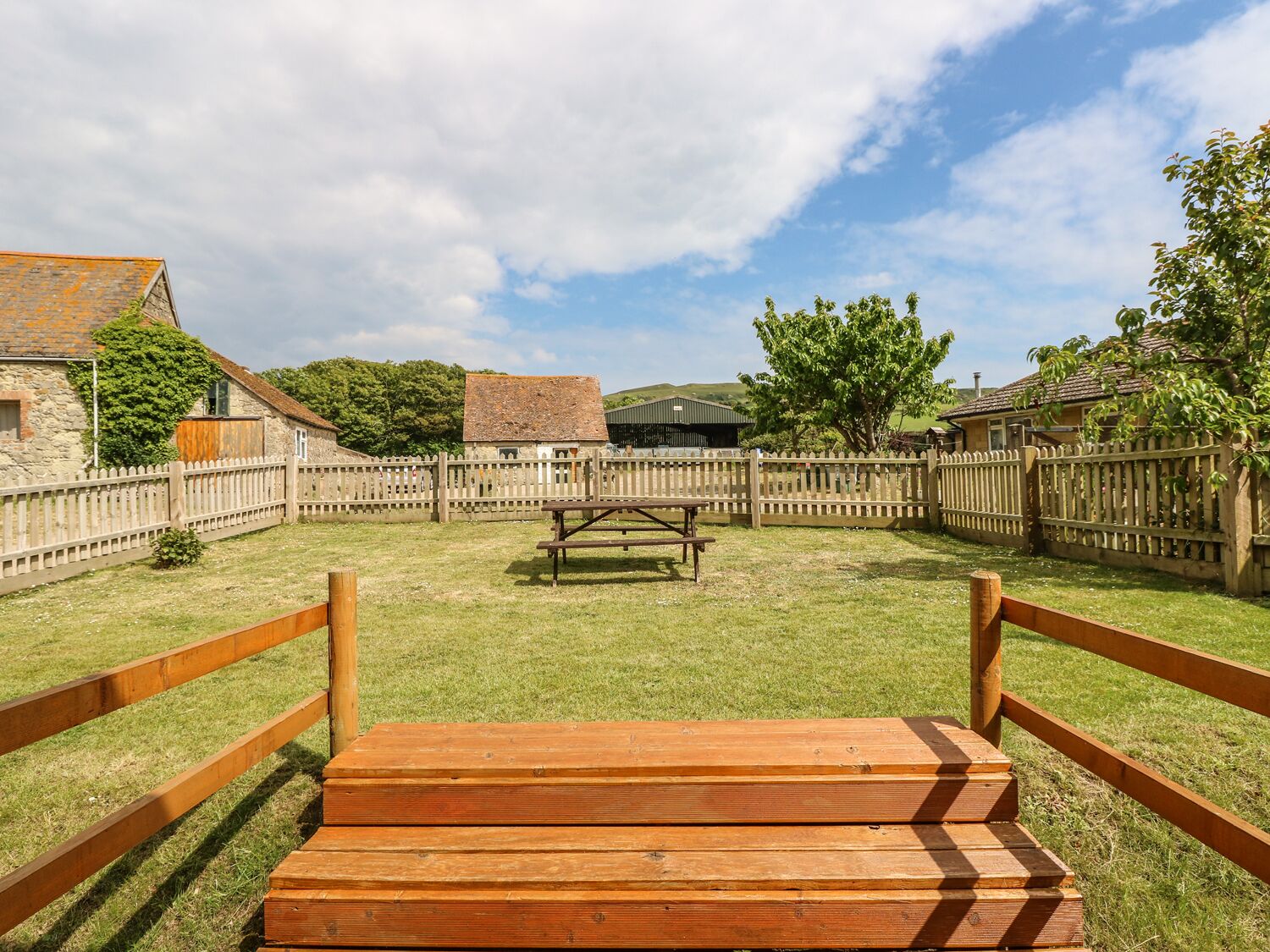 A grassy outdoor area with a wooden bench and picnic table, surrounded by a wooden fence and stone buildings.