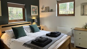 Een slaapkamer met een bed, twee ramen, een nachtkastje, een lamp en een dressoir.