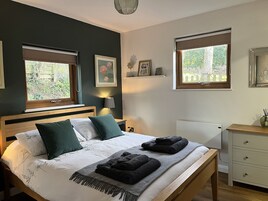 Un dormitorio con una cama, dos ventanas, una mesita de noche, una lámpara y un aparador.