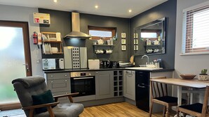 Een moderne keuken met grijze kastjes, een inbouwoven, een magnetron en een koffieapparaat. Er is een eettafel met stoelen en een stoel met kussen.