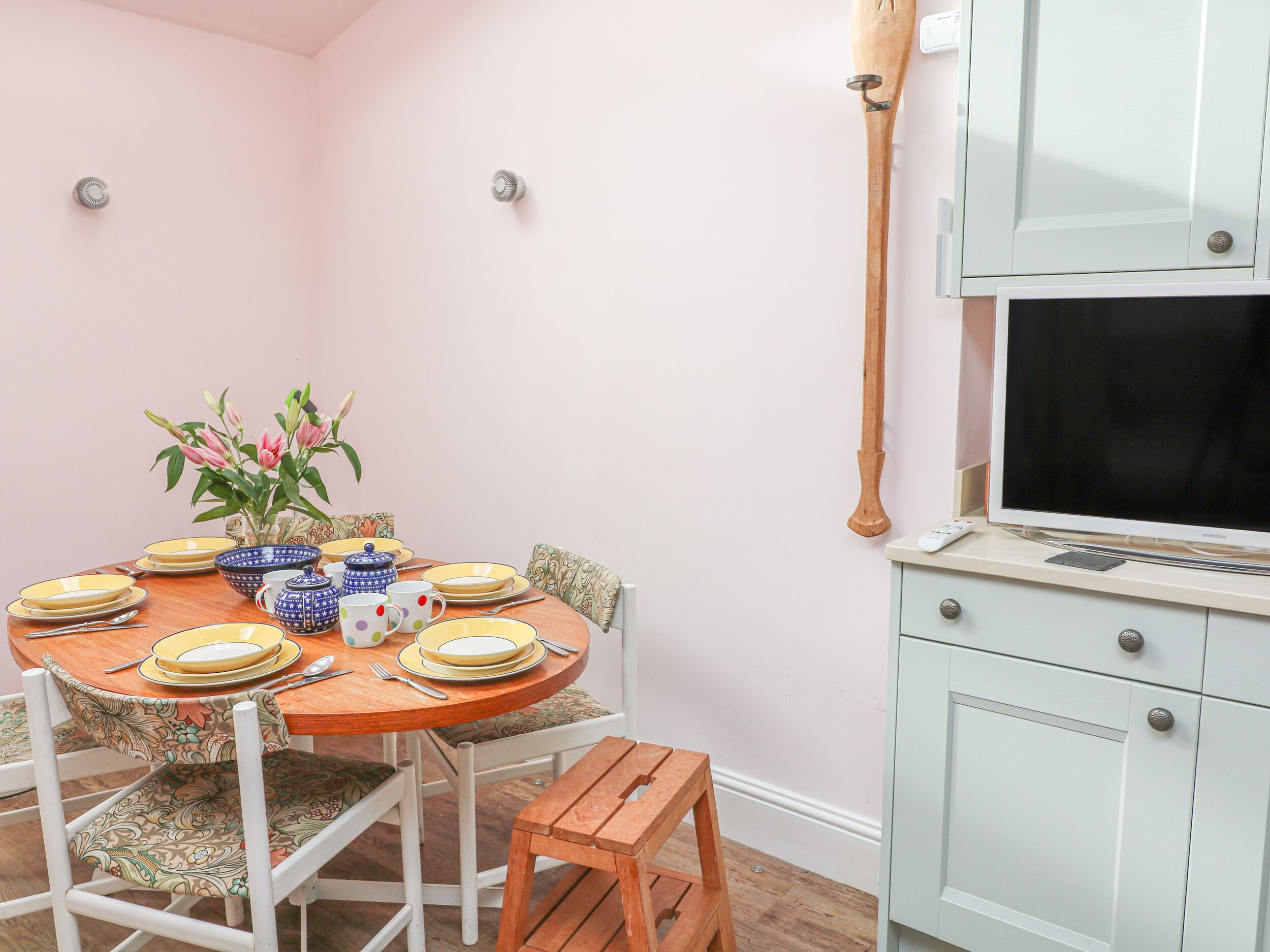 Uma sala de jantar com uma mesa de madeira posta para quatro pessoas, rodeada por cadeiras brancas com almofadas estampadas. Há um vaso com flores sobre a mesa, e uma televisão fixada na parede à direita.