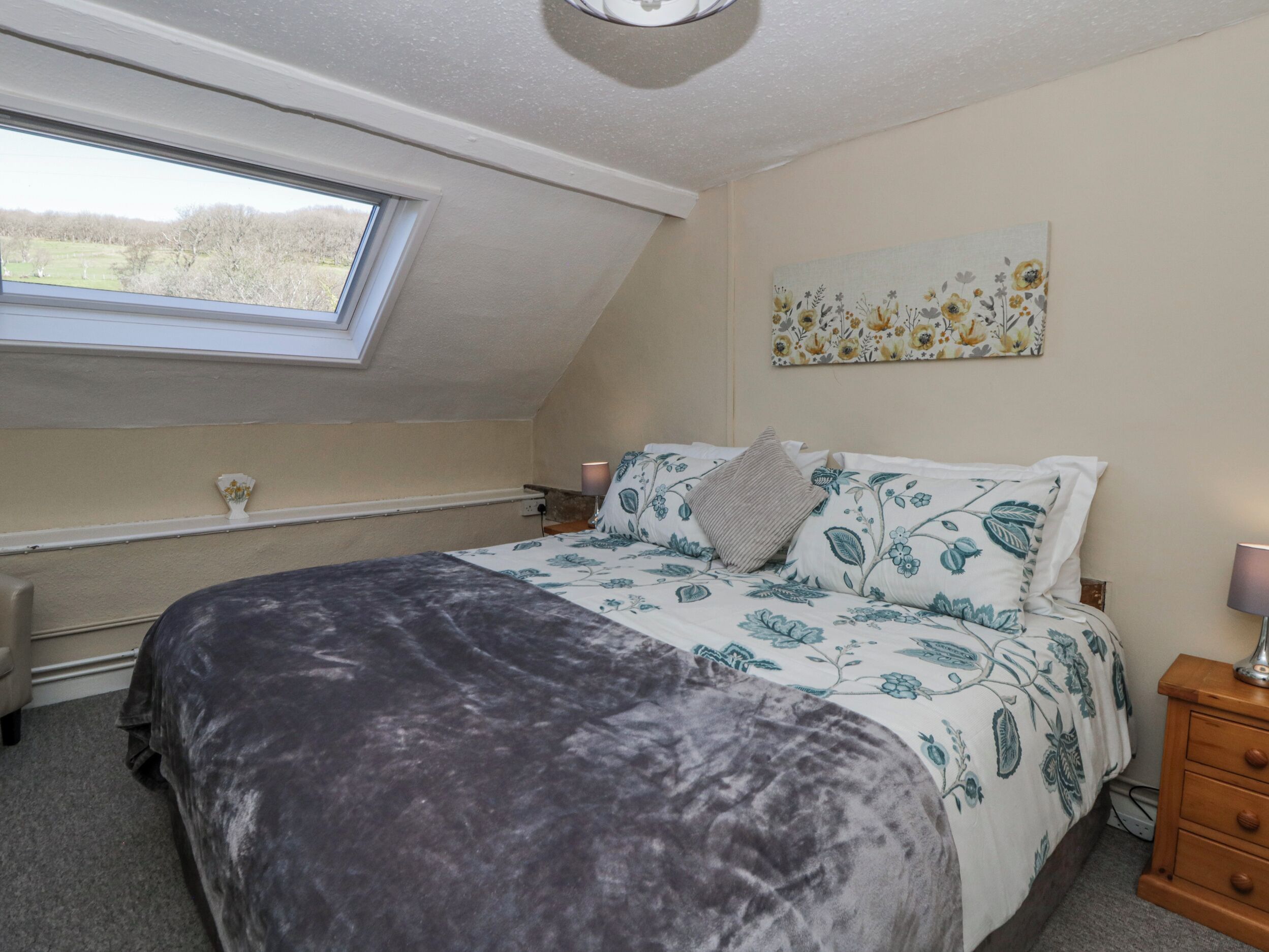 A bedroom with a skylight, a bed with floral bedding, a bedside table, and a wall-mounted painting.