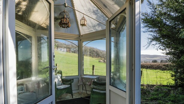 A conservatory with a view of a green field, a wicker chair, and a small table. - Penlone Cottage (Rhayader)