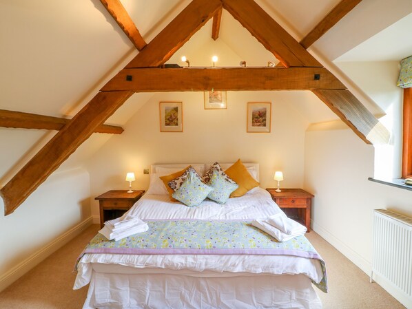 A bedroom with a sloped ceiling, wooden beams, a bed with multiple pillows, two bedside tables with lamps, and a radiator. - Great Bradley Cottage (Minehead)