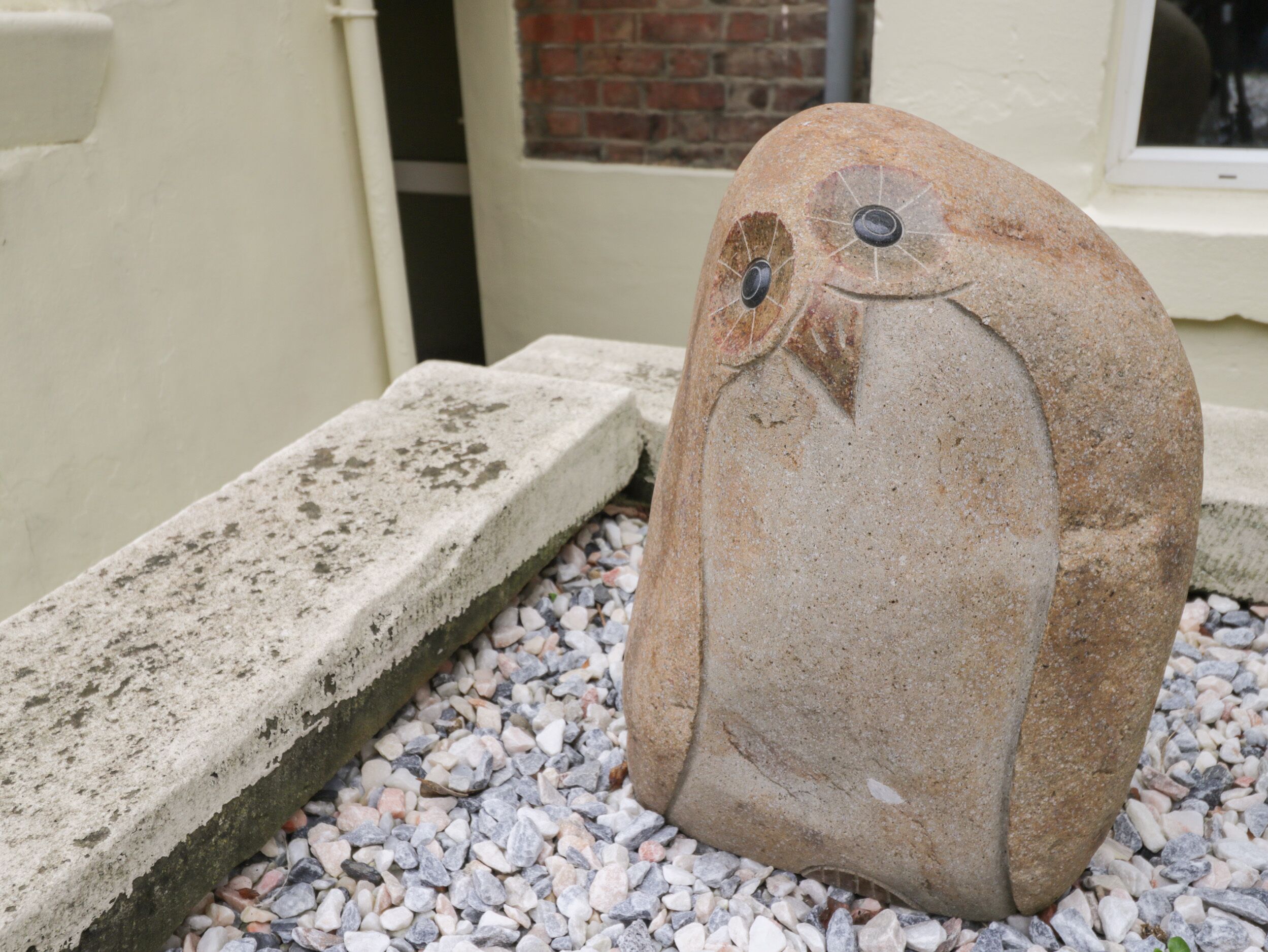 A stone sculpture of a bird with a hole for an eye and a small hole for an ear, placed on a gravel surface next to a concrete ledge.