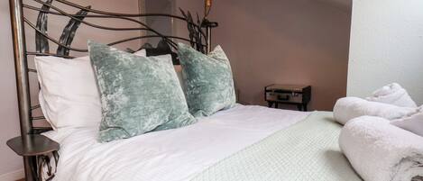 A neatly made bed with a white duvet and two green pillows, a small bedside table with a lamp, and a suitcase on the wall.