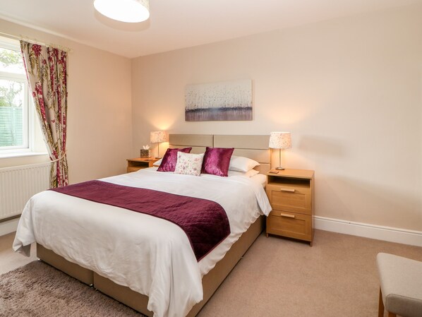 A bedroom with a bed, two bedside tables, a nightstand, a window with curtains, a radiator, and a wall-mounted artwork. - Berrymoor Cottage (Brampton)