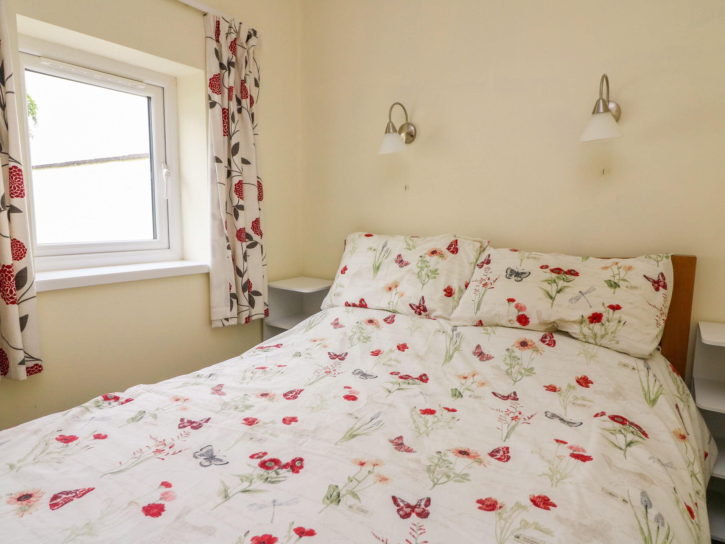 Un dormitorio con una colcha de flores, una ventana con cortinas y dos lámparas fijadas a la pared.