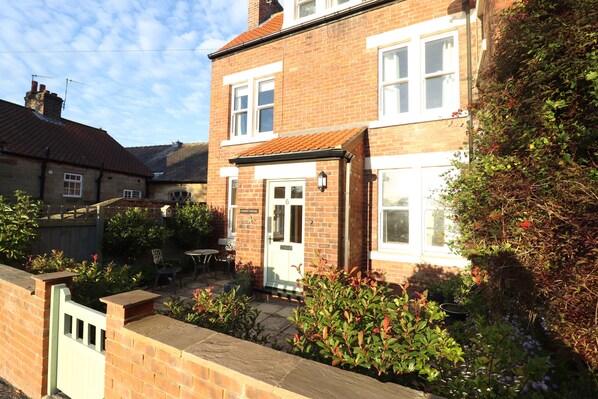 A two-story brick house with a front door, a small patio with a table and chairs, and a garden with various plants. - Arden House (Whitby)