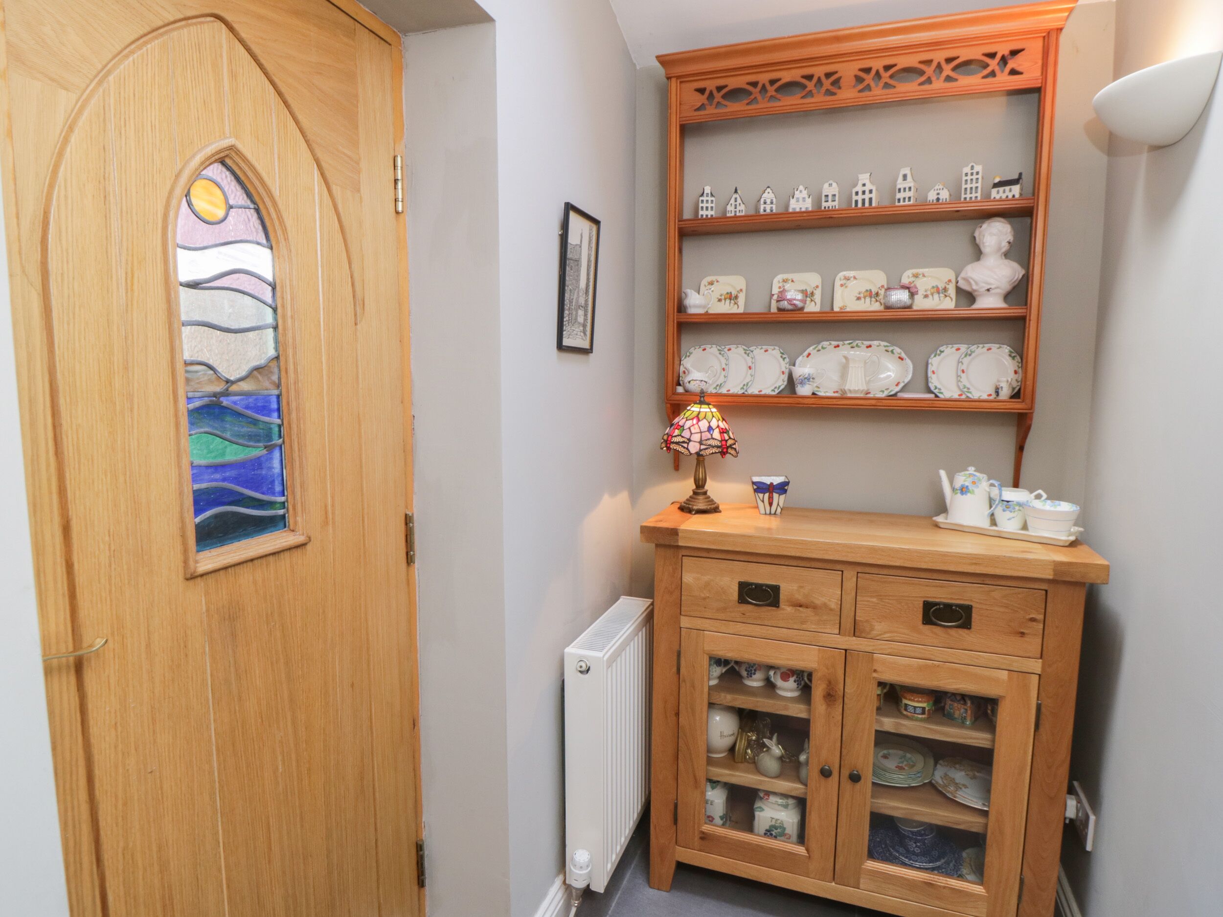 Une porte en bois ornée d’un vitrail, un buffet en bois avec des étagères remplies d’assiettes et de bols, et un radiateur.