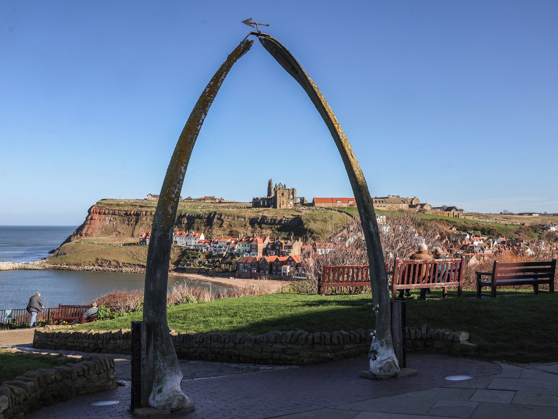 A coastal town with a prominent church and castle on a hill, a clear sky, and a park with benches.