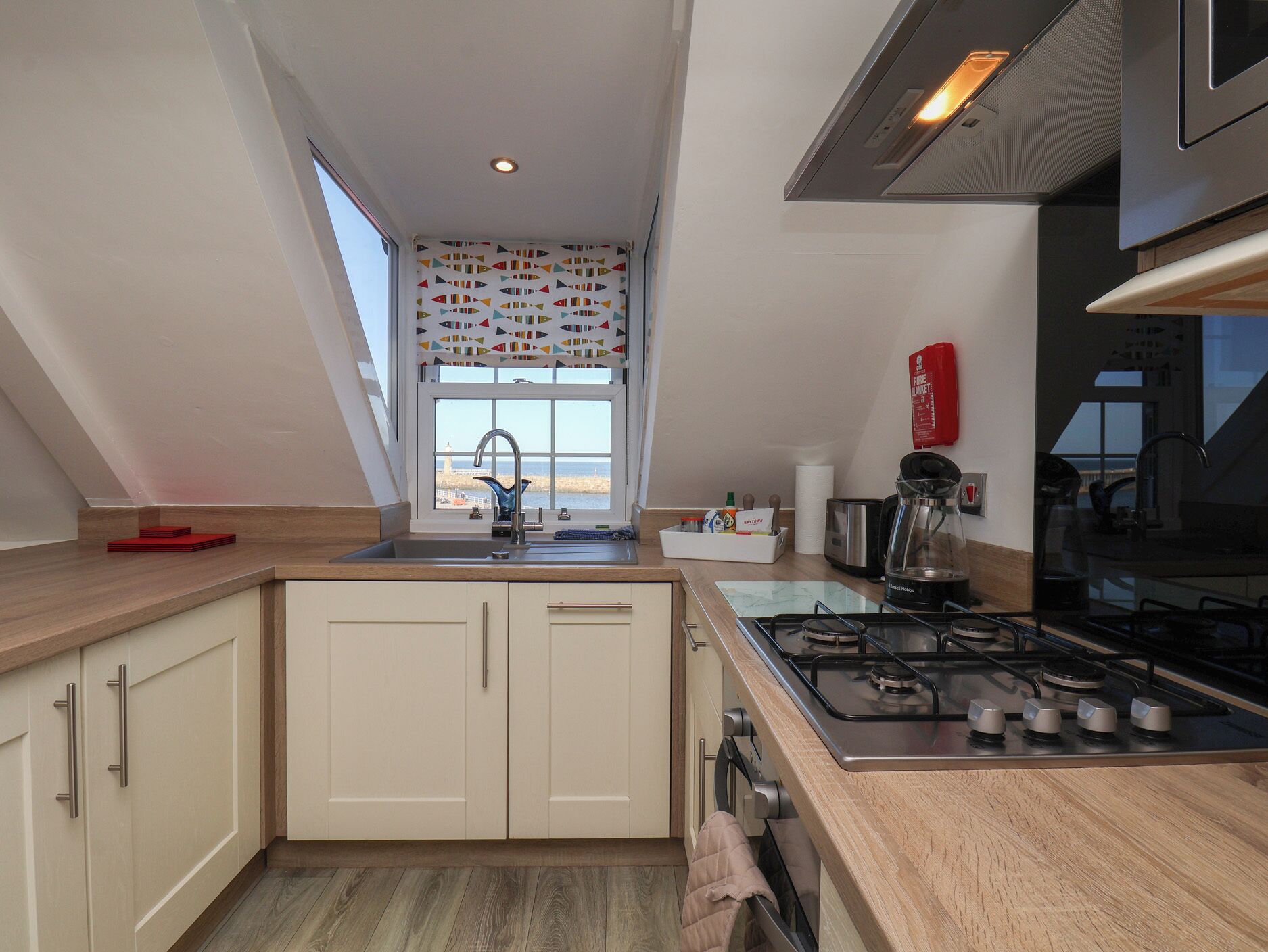 A modern kitchen with a view of the sea, a gas stove, and a window with a geometric curtain.