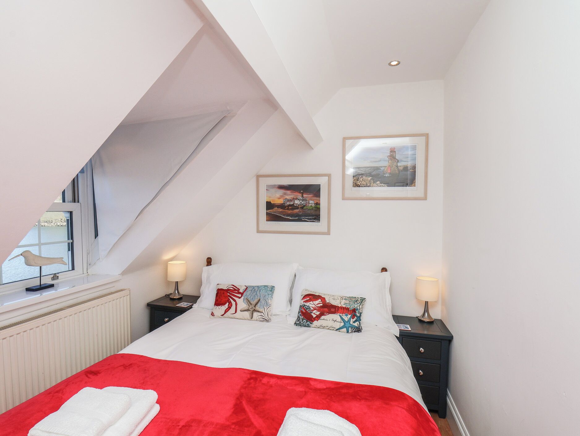 A bedroom with a sloped ceiling, a bed with a red blanket, two nightstands with lamps, and two framed pictures on the wall.