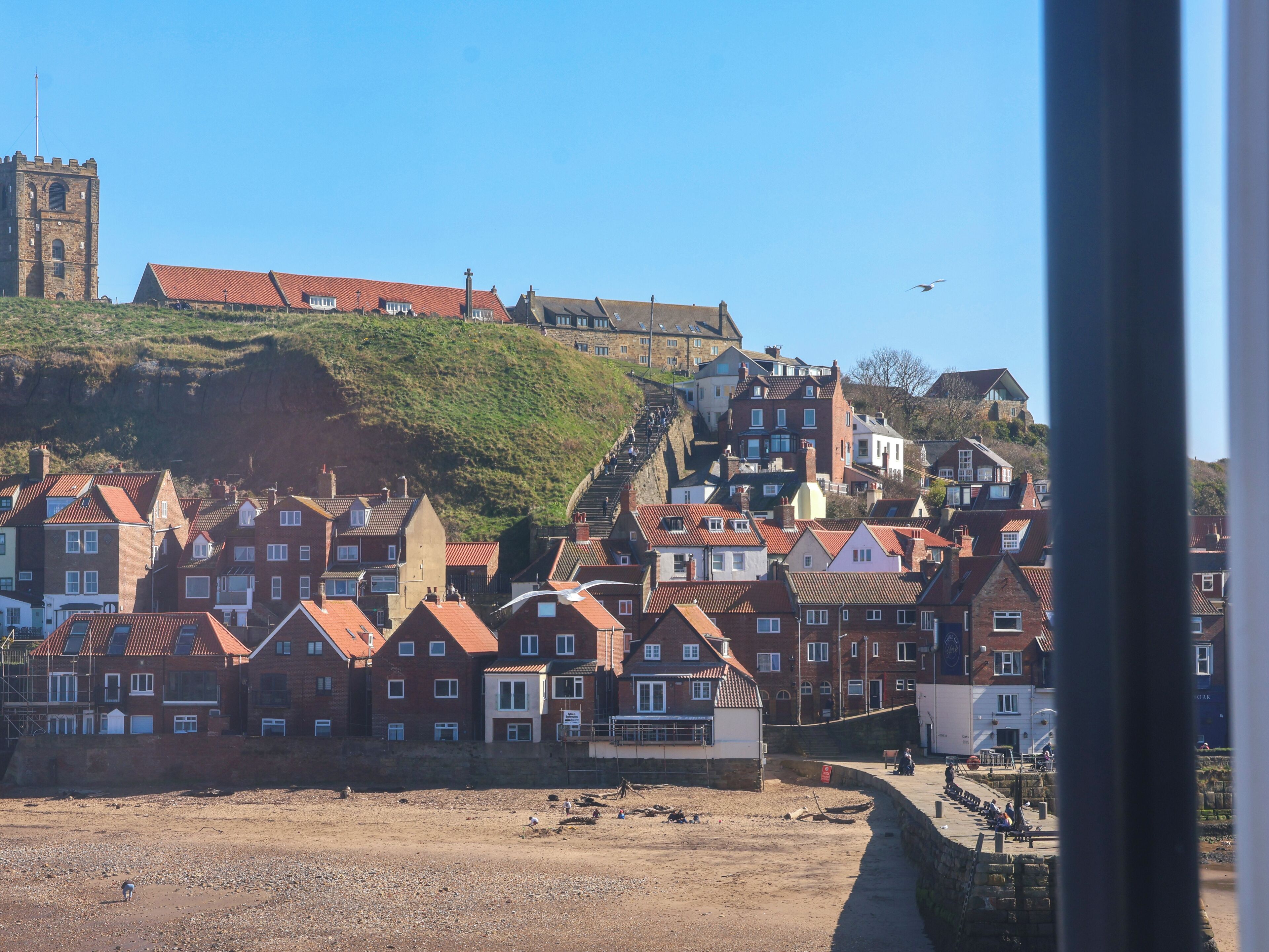 A coastal town with historic buildings, a prominent church tower, and a hill with a castle-like structure.