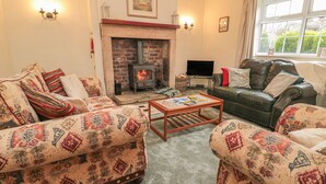 A cozy living room with patterned sofas, a fireplace, a television, and a window with curtains. - Wordsworth Cottage West (Morpeth)