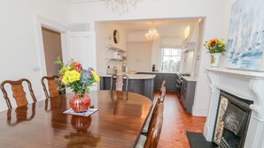 A dining room with a wooden table and chairs, a vase of flowers, and a kitchen in the background. - Mor Awel (Beaumaris)