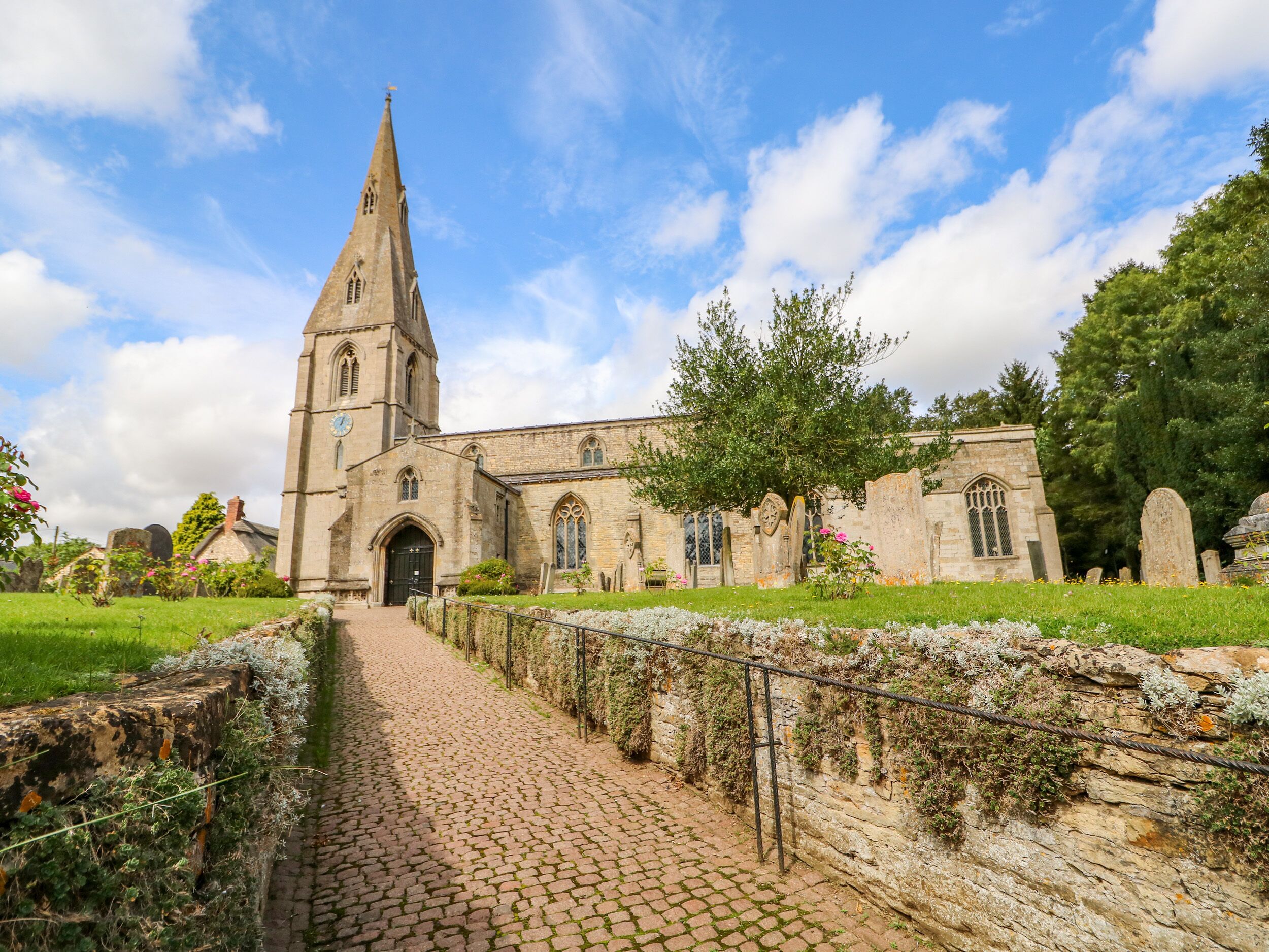 A historic church with a tall spire, a paved pathway leading to the entrance, and a stone wall with greenery.