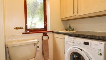 Une petite salle de bain comprenant des toilettes, un lave-linge, un lavabo et une fenêtre donnant sur un espace verdoyant.