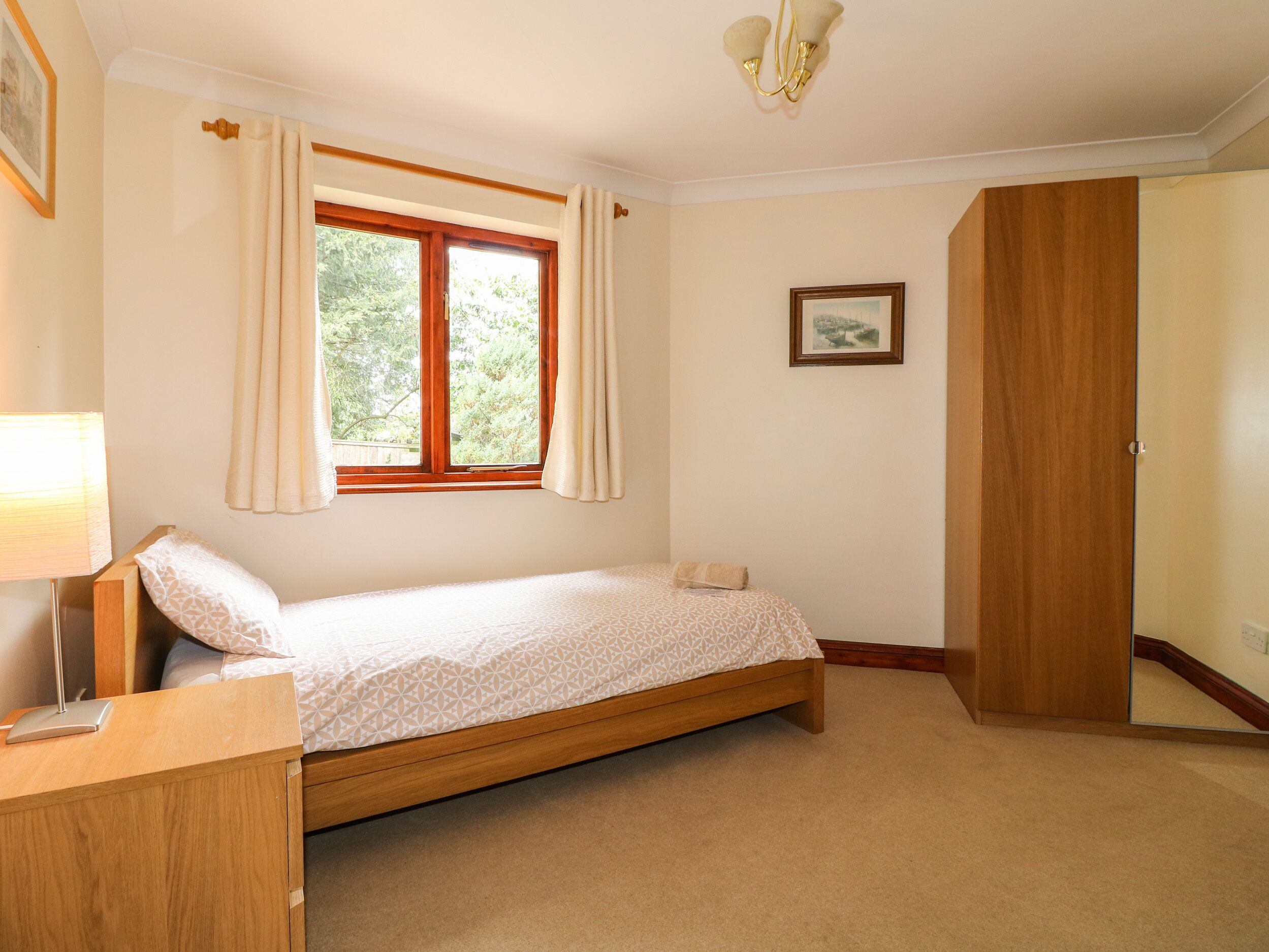 Quarto com uma cama de solteiro, mesa de cabeceira de madeira, escrivaninha, janela com cortinas, quadro emoldurado na parede e guarda-roupa.