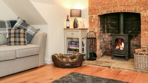 A cozy living room with a brick fireplace, a sofa with plaid cushions, a dog bed, and a small shelf with books and a wine bottle. - Tilly's Cottage (Westbury)