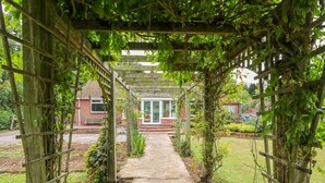 A garden pathway under a wooden pergola with climbing plants, leading to a building with a red brick facade and a white door. - Glynde (Dover)