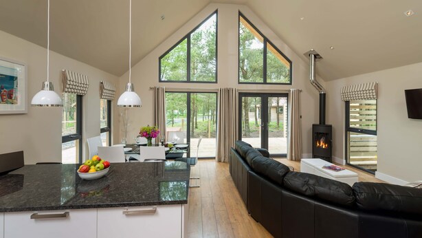 A spacious living area with a large black leather sofa, a dining table with chairs, a kitchen island with a fruit bowl, and a fireplace. - Bracken Howe Lodge (Whitby)
