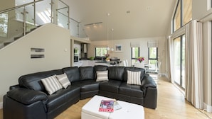 A modern living room with a black leather sectional sofa, a white coffee table, and a staircase with glass railings leading to an upper level. - Bracken Howe Lodge (Whitby)