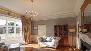A spacious living room with a large window, a chandelier, a fireplace, and a classic wooden cabinet. - 12 St Hilda's (Whitby)