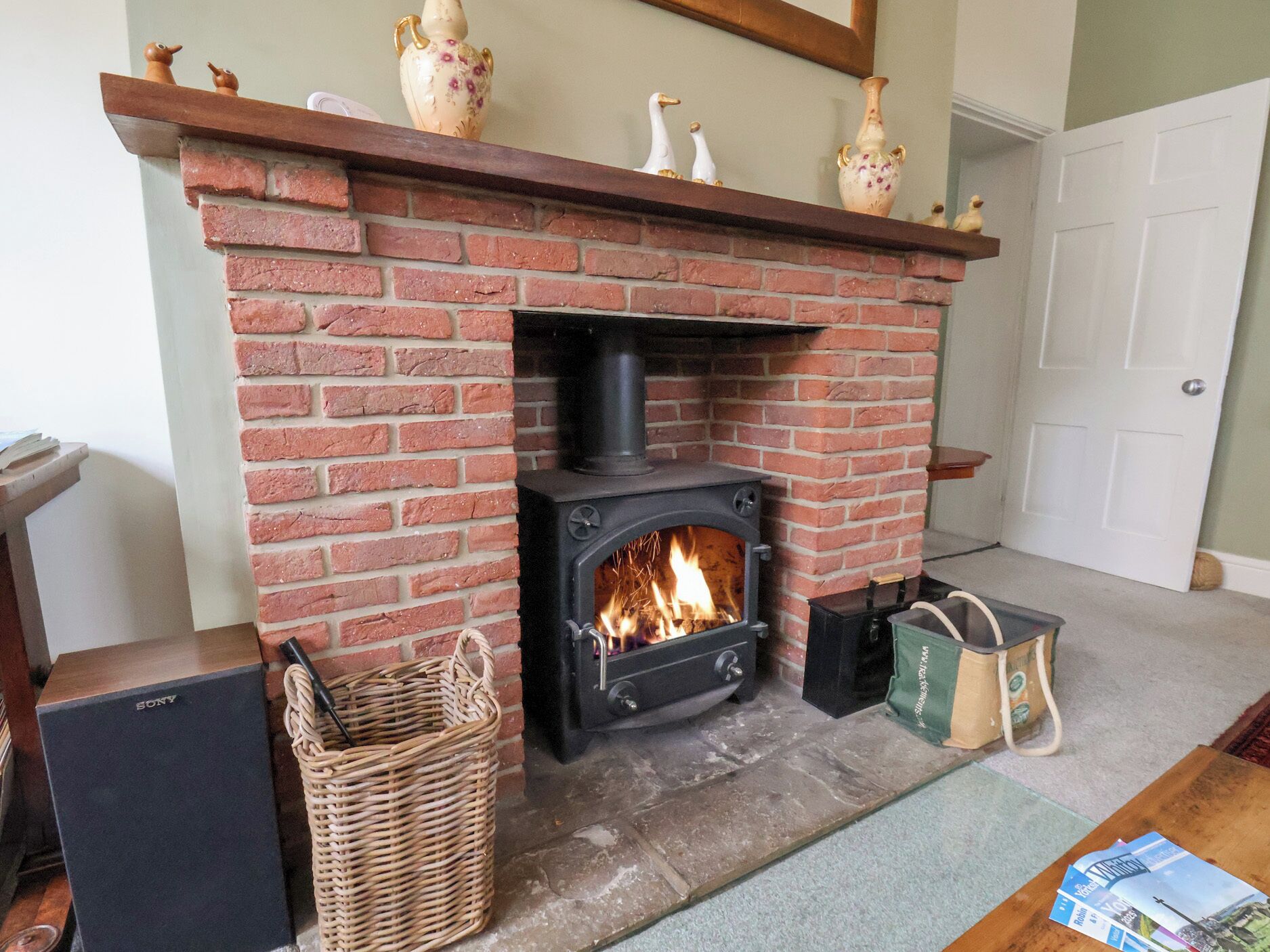A brick fireplace with a black wood stove, a woven basket, a green bag, and a Sony speaker.