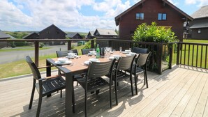 Une terrasse en bois avec une table dressée pour le repas, d’où l’on peut admirer une rangée de maisons et un ciel dégagé.