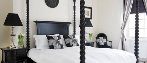 A four-poster bed with a dark headboard, white bedding, and decorative pillows. There are two bedside tables with lamps and a vase of flowers. A framed picture hangs on the wall above the bed.