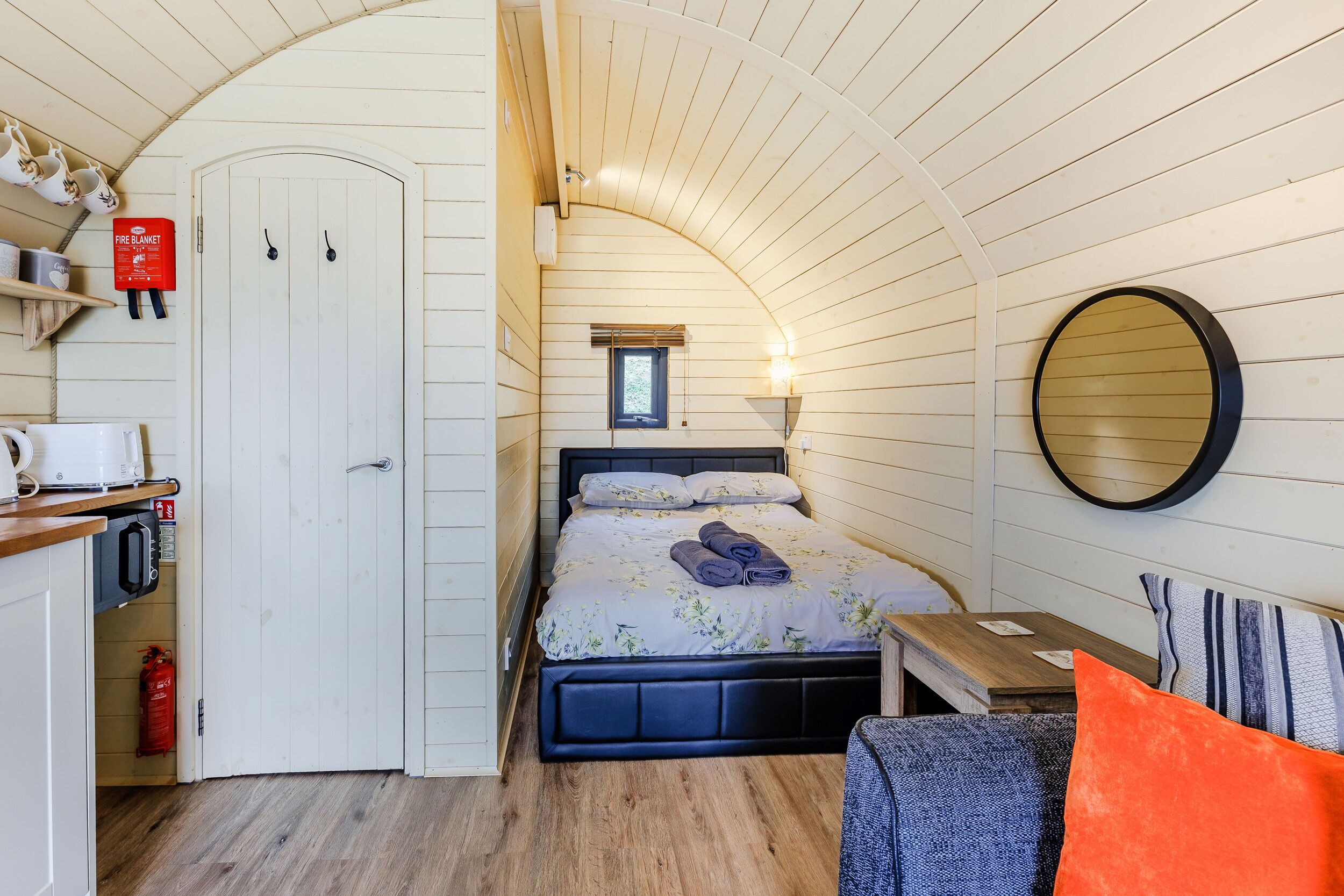 A cozy room with a bed, a small table, a chair, a round mirror, and a window.