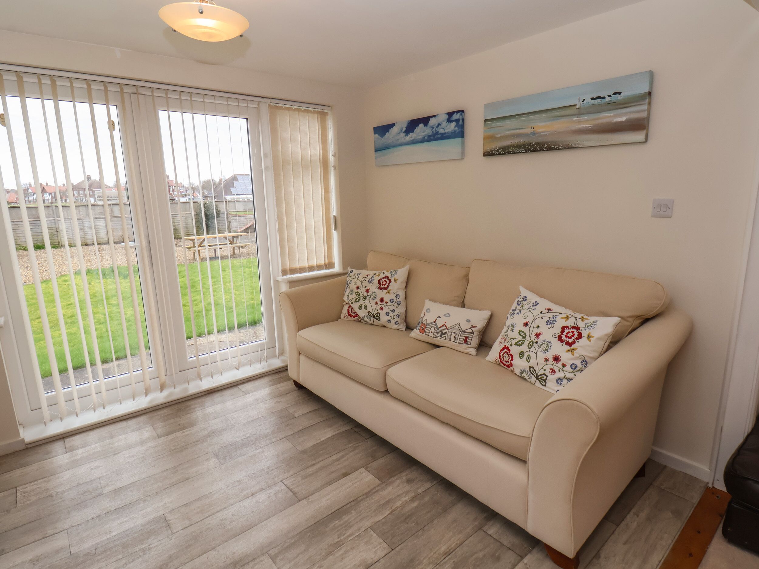 A beige sofa with floral cushions, a wooden floor, a sliding glass door leading to a garden, and two landscape paintings on the wall.