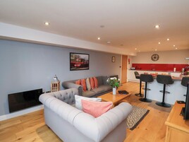 Una sala moderna con un sofá gris, una mesita con un jarrón de flores, y una zona de cocina con detalles en rojo y taburetes de bar.