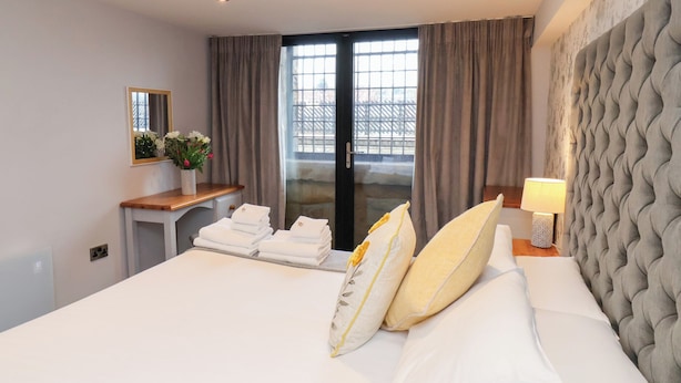 Quarto de hotel com cama, escrivaninha com toalhas, espelho, janela com cortinas e parede com padrão texturizado.