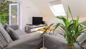 En moderne stue med en grå sofa, et sofabord og et fjernsyn. Der er et stort vindue med udsigt til grønne omgivelser.