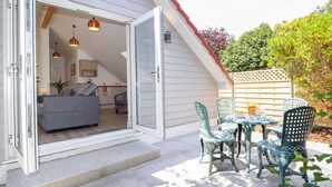 En terrasse med et bord og stole, samt en skydedør der fører ind til en stue med en sofa og en lampe.