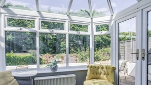 Een oranjerie met uitzicht op een tuin, een gele fauteuil, een radiator en een tafel met een plant.